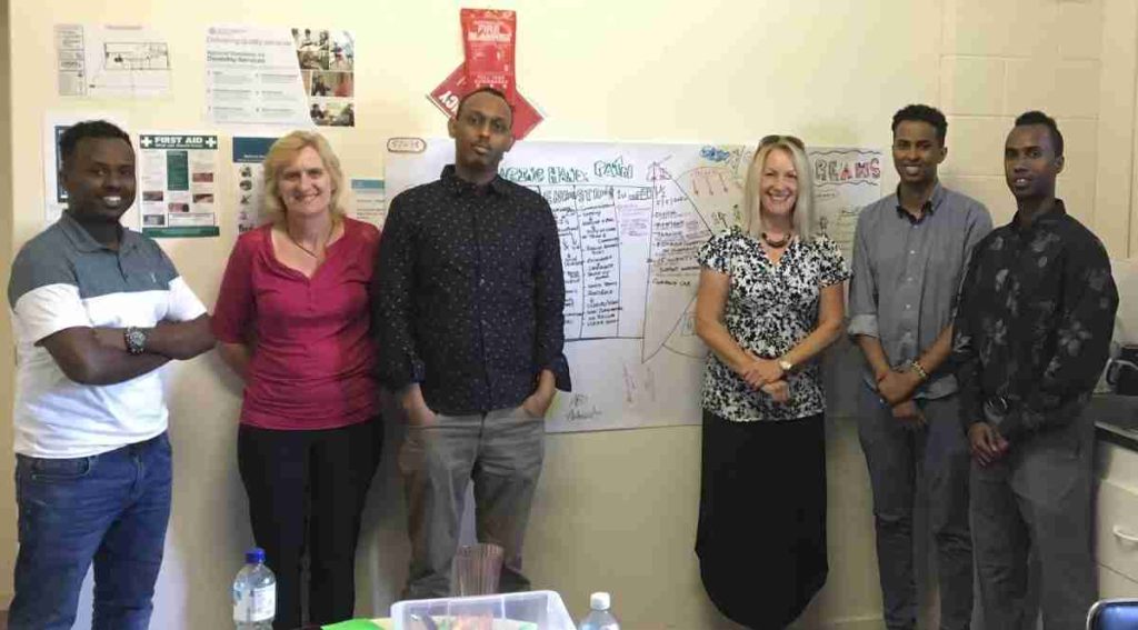 2 women and 4 men stand in front of a cream coloured wall on which a graphical PATH plan is hanging.