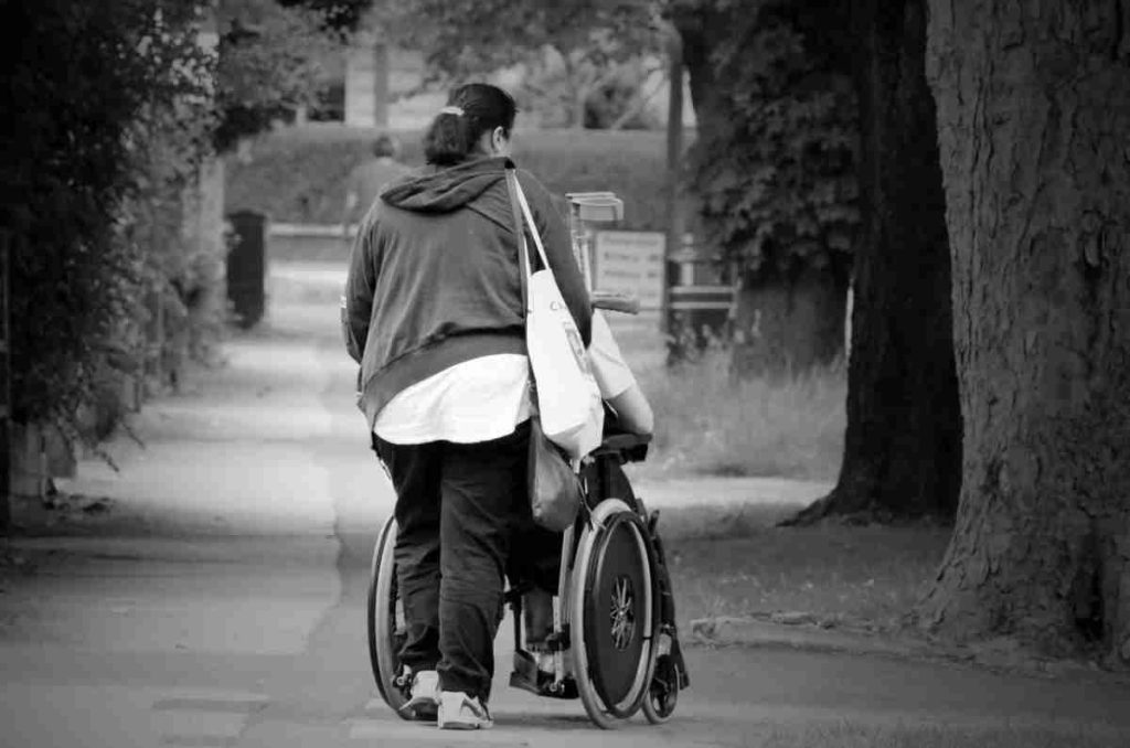 Black and white photo of the back of a woman pushing a person in a wheelchair.