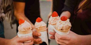 A close up of four hands, each holding a strawberry ice cream cone with a fresh strawberry on top.