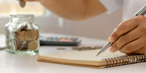 Image of a person dropping a coin into a jar while using the other hand to write in a notebook