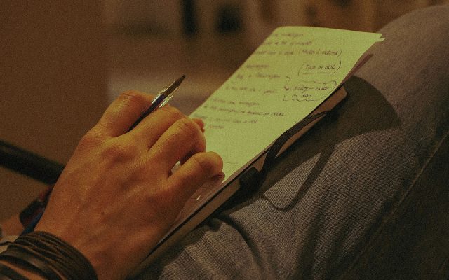 Hand holding pen with notepaper resting on leg